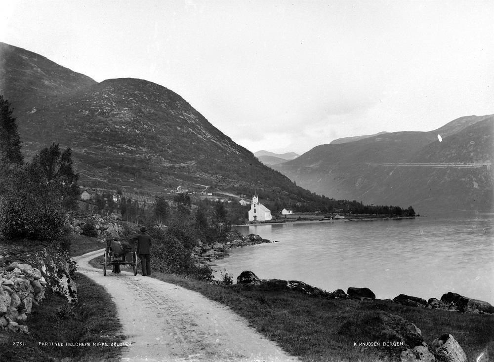 Postvegen ved Jølstravatnet kring 1890 med utsyn nordover til Helgheim kyrkje. Fylkesmann Aubert sa i 1854: 'Fulstændiggjørelsen af den Communicationslinie, der skal sætte Districtets forskjellige Hoveddele i Forbindelse med hinanden maa jeg ansee som en Sag af første Rang naar Spørsmaalet er om Bevilgelse af Midler til Veiveæsenets Fremme.' Likevel vart vegen bygd smalare (3,8 m i staden for 5 m). Den utbetra vegen til dampskipsstoppestaden Steinen i Førde vart frå 1859 svært viktig for bygdene i J...