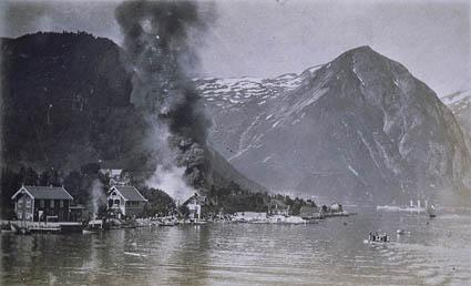 Brannen i Balestrand i 1911. Tjukk og svart røyk veltar opp frå brannstaden. Biletet må vera teke etter at brannen hadde herja ei tid. Like framføre brannstaden ser me huset til Kristen Virkesdal som vart berga. I fjøra nedanfor brannstaden er det mykje folk som tek del i sløkkinga. Dei to husa til venstre i biletet er Kai-huset og Cooperhuset. Desse står i dag.