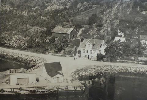 Bryggja på båtstoppestaden Slinde (Lunden), kaihusa og våningshuset år 1955. John Lidal sette bryggja i stand då han flytte til Lunden i 1910. Dei brukte for det meste furestokkar. Til høgre for kaiskuret var sett ned ein stor stokk som festepåle. Den var bardunert i land. Båtane la til med baugen inn mot land. Det var også båtfeste i andre enden av byggja.