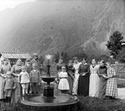 Eigaren av garden Rikheim i Lærdal, Claus Søfren Ruhmor (1850-1915) (midten) med familie på garden.
Kona Inga Eilertsen (1857-1949) med borna. Truleg er det frå venstre framme: Alette Marie Rumohr (1879-1962), Johan (1887-1965), Claus Søfren (1889-1927) og Fredrik Johan Rumohr (1882-1945). Jenta til høgre for fontena i kvit kjole, er truleg Marianne Rumohr (1885-1970). Heilt til høgre truleg Claudine Rumohr (1880-1967). Kvinna til høgre for fontena er Dagny Rumohr (?-1898). Kvinna i ljos, blomstrut ...