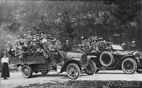 Bilen til venstre er ein Opel med Olai Farsund som sjåfør, medan sjåfør Samuel S. Veiteberg køyrer den andre bilen, ein Adler. Biletet er teke på Kleivedammen på Sandane rundt 1920. Opelen er truleg den første bilen til A/S Fjordenes Automobilselskap(sjå ovanfor). Ved turkøyring vart lasteplan og bilsete ofte fylte heilt opp. Bilane har kompakte gummihjul utan fjøring. Ein tur på lasteplanet var difor ikkje særleg behageleg. Frå 1930 vart det kravd luftdekk på alle nye lastebilar. 

 