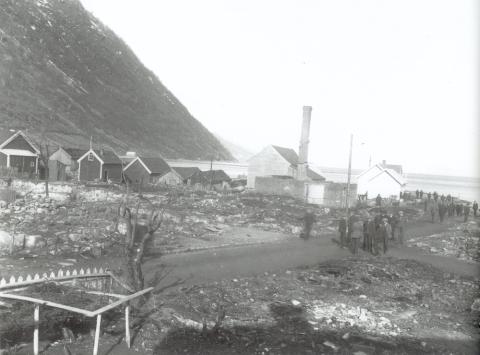 Slik såg det ut vest for hovudgata etter brannen i 1922. Ruinane etter Forbruksforeningen sitt bakeri nærast kaiområdet. Hus og naust mot vest vart ikkje råka av brannen. Mykje folk er ute og diskuterer det som har skjedd.