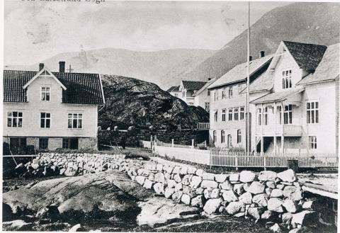 Bak det  kvite huset til høgre ser me bustadhuset og verkstaden som Bjørn Sande bygde opp etter at fyrste verkstaden hans brann i 1911. Her har det vore verkstad, saftpresseri, vevstove, sal av souvenirar  og kafe.  Huset til høgre framme i biletet er doktorbustaden som brann ned  i 1950-åra. Til venstre i biletet ser me huset til Virkesdal som vart berga frå brannen takka vera innsatsen frå engelske og tyske sjømenn. Me ser og korleis sjøen den gongen gjekk like opp til kjellaren på dette huset. I ...