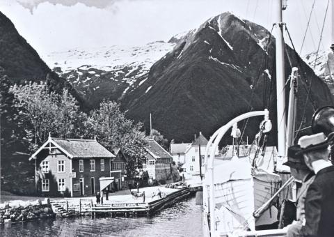 Slik har mange reisande møtt Balestrand når dei har kome med båt inn fjorden. Her kjem D/S <i>Kommandøren</i> til kai i Balestrand, truleg på utgåande rute kl. 12 på dagen, ein sommardag i 1936. <i>Kommandøren</i> gjekk i rute mellom Bergen og Indre Sogn. Her ser vi bryggja som vart  bygt i 1911 og Kaihuset. Ved sida av kan ein så vidt skimte bygningen Bom Bom før Cooperhuset.